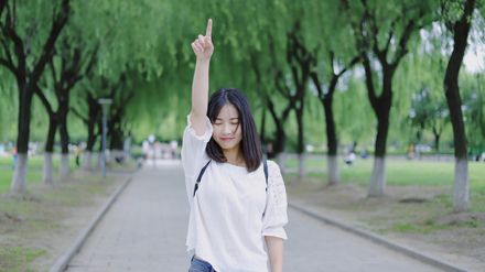 Jeune femme les yeux fermés et souriante, pointant le doigt vers le ciel sur fond de parc