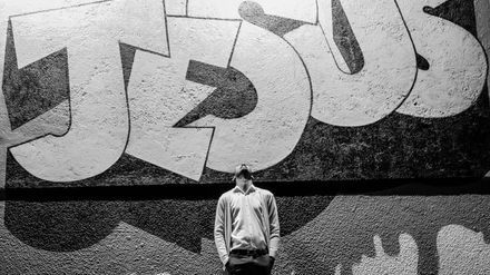 Photo en noir & blanc d'un homme le visage au ciel devant un mur où se trouve un immense tag "Jésus"